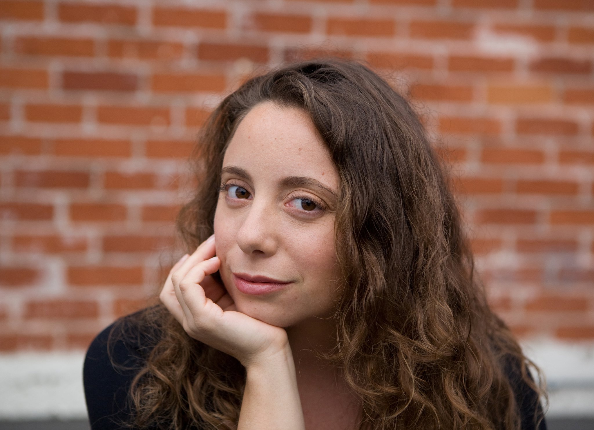 Woman with brown wavy hair and hand on chin against backdrop of brick wall